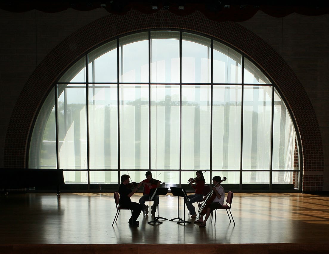 Silhouetted string quartet rehearsing in a sunlit hall with a large arched window behind them.